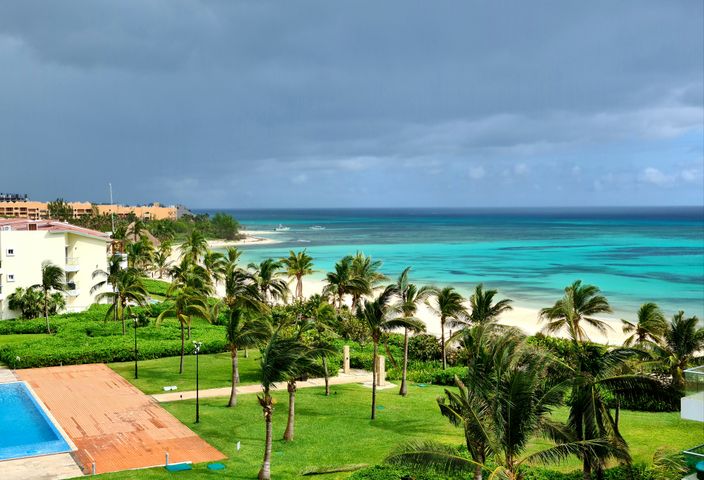 Aerial view of a tropical beach resort with lush palms, turquoise ocean, and a swimming pool under a cloudy sky.