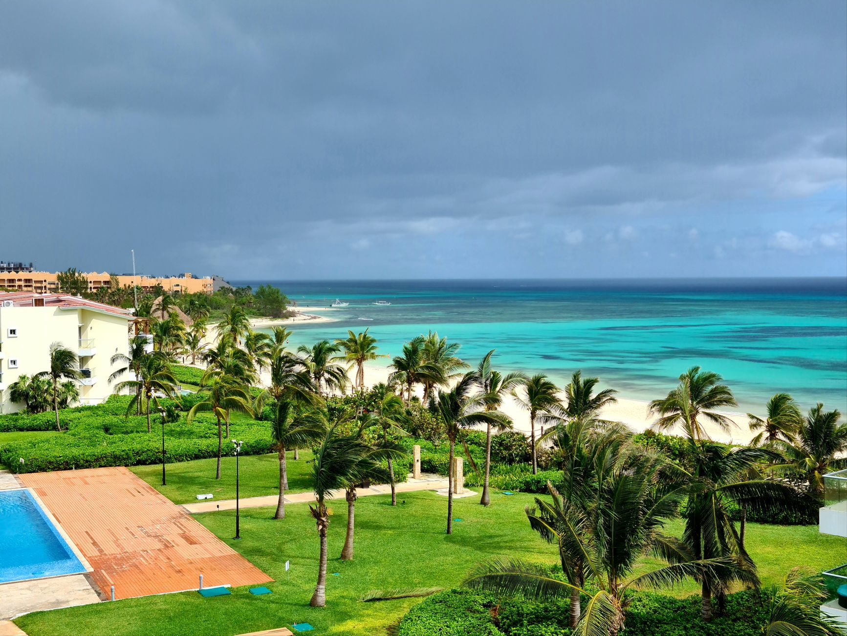 Aerial view of a tropical beach resort with lush palms, turquoise ocean, and a swimming pool under a cloudy sky.