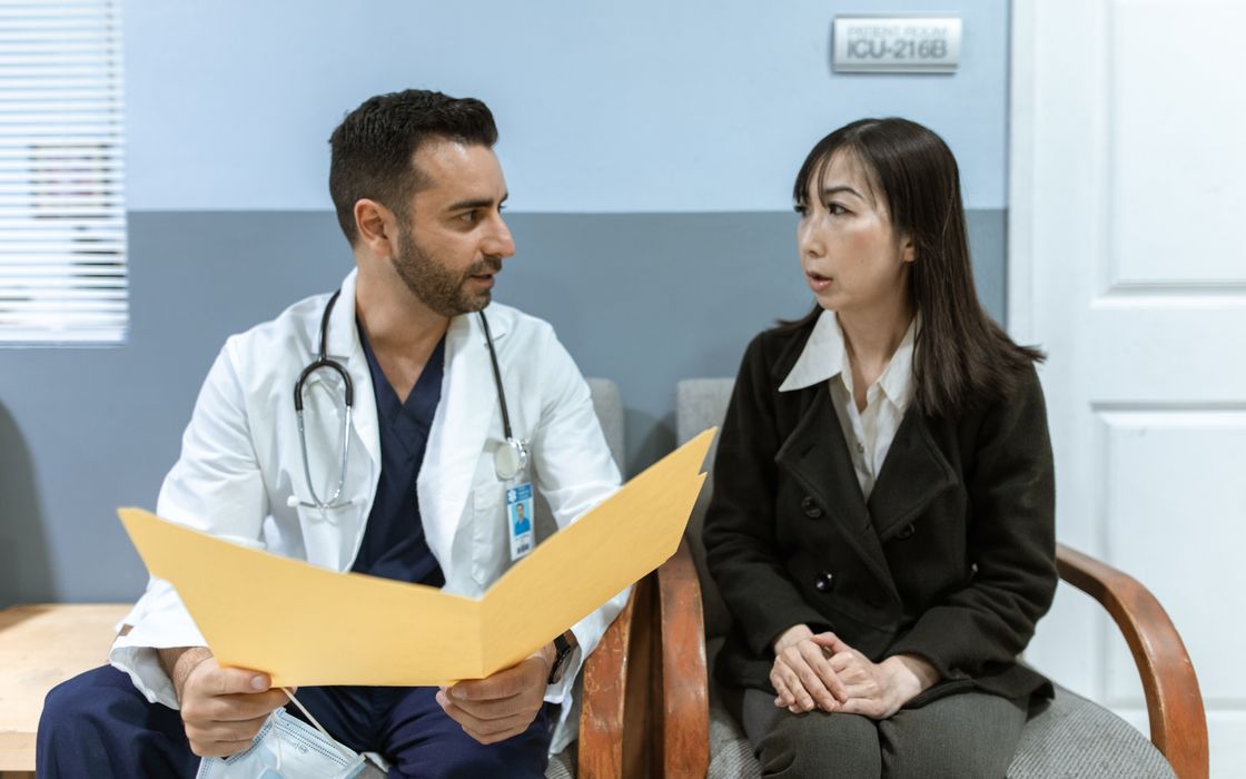 A doctor holding a folder of records talks with a female patient during a consultation.