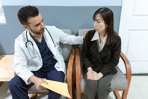 A doctor in scrubs and white coat comforting a patient sitting in a medical office.