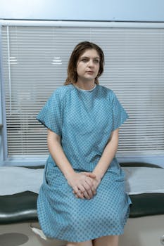A woman in a hospital gown sitting on an exam table, looking contemplative.