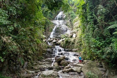 Free Back view full body of anonymous male near fast waterfall streaming through rocky hill near green plants in summer day Stock Photo