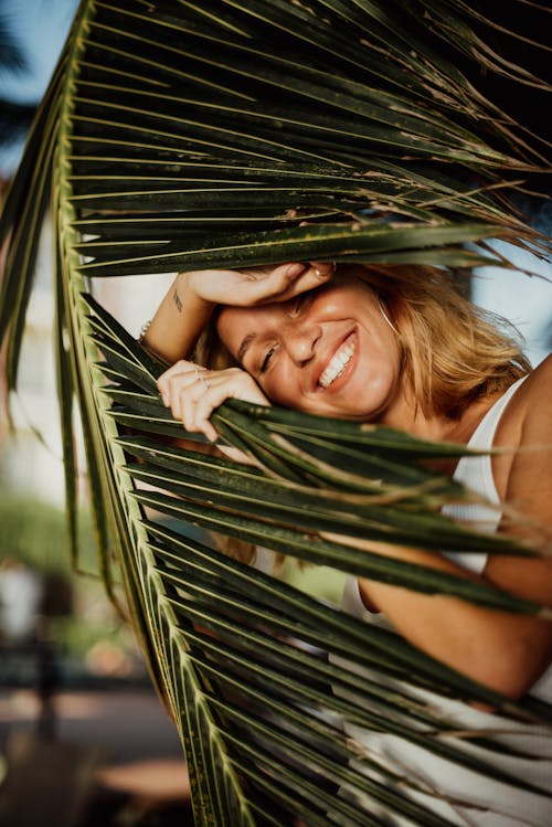 Free Woman in White Tank Top Behind Palm Leaves Smiling Stock Photo