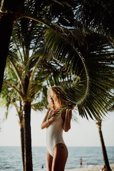 Blonde woman in one-piece swimsuit posing by palm trees on a sunny Bali beach.
