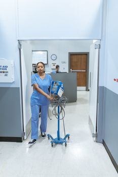 A nurse in blue scrubs walks through a hospital corridor with medical equipment.