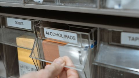 Detail of a hand retrieving alcohol pads from a medical supply dispenser on a shelf.