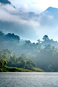 Landscape of pond near green shore with grass and trees on hills with plants under cloudy sky in sunny summer day