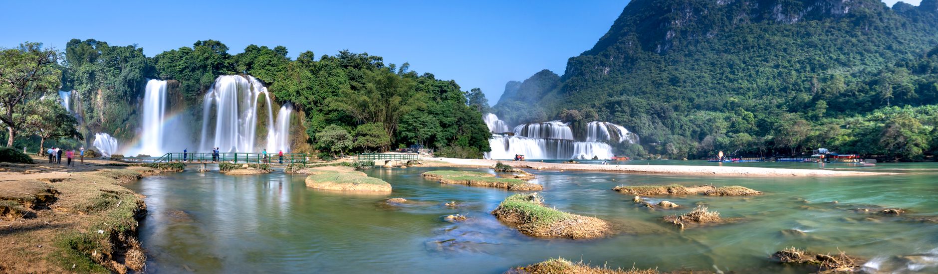 Landscape of pond with waterfalls near rocky shore with grass and trees on mountains with plants under blue cloudless sk