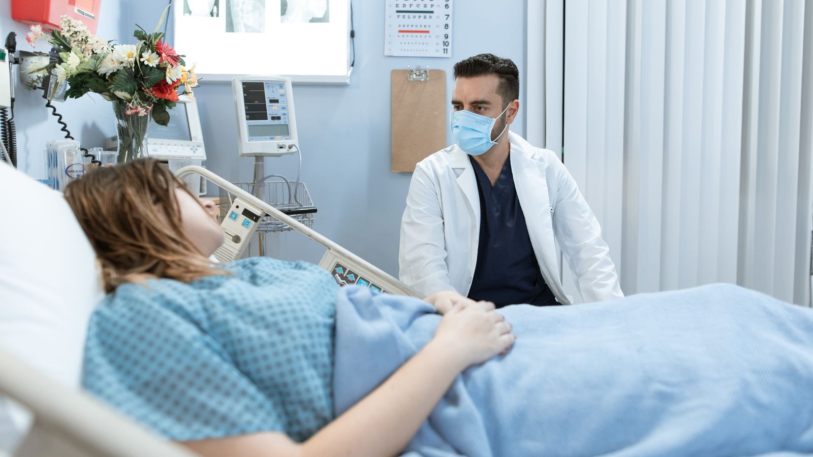 A doctor in a face mask talks to a patient in a hospital bed, providing care and consultation.