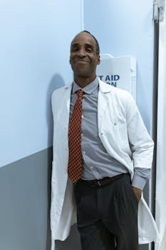 A confident black male doctor stands smiling in a hospital corridor wearing a white coat.