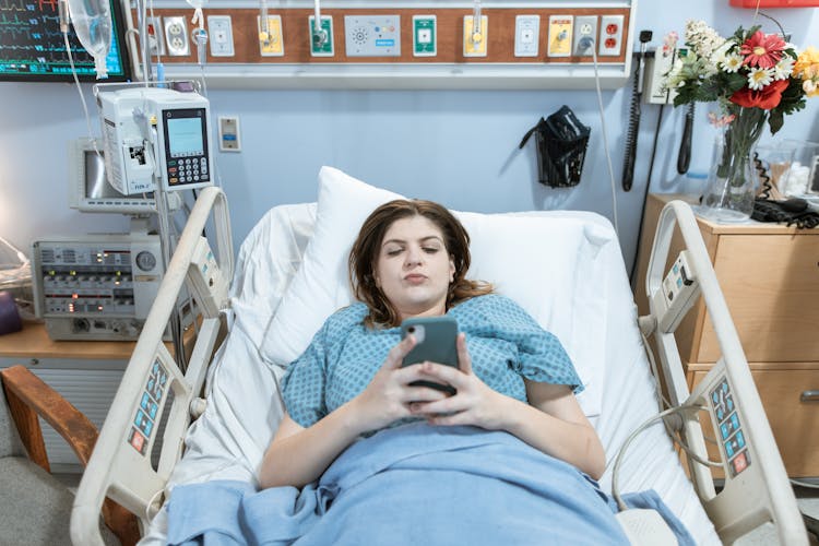 Female Patient Using Phone While Lying On A Hospital Bed 