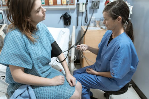 A nurse in blue scrubs checks the blood pressure of a female patient in a hospital room.