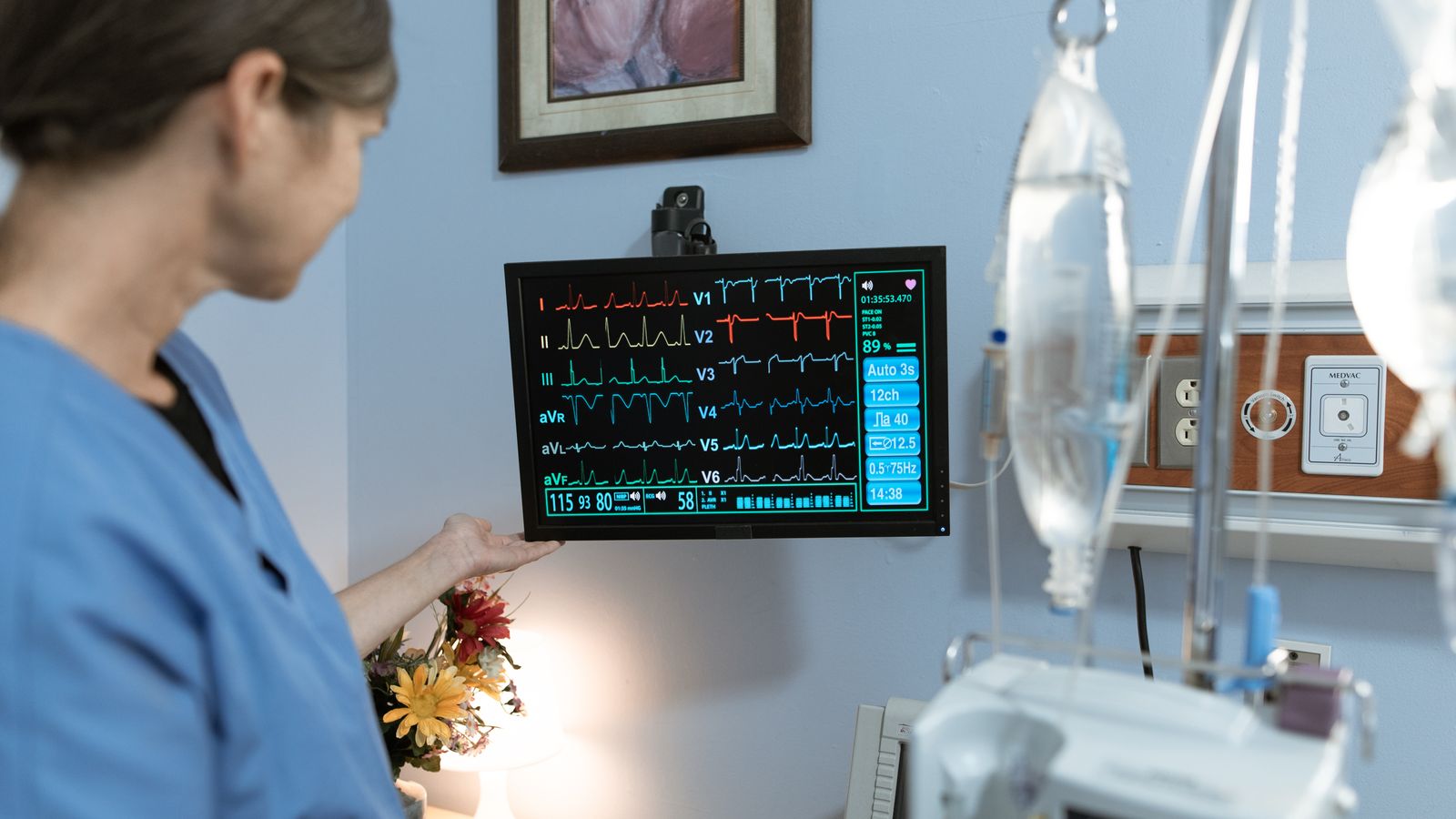 A nurse in blue scrubs examines a medical monitor displaying vital signs in a hospital setting.