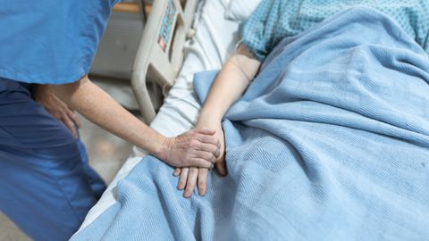 A nurse in scrubs offers comfort by holding a patient's hand in a hospital bed.
