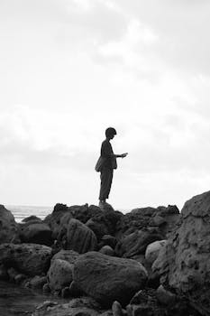 Black and white image of an individual standing on coastal rocks, looking contemplative.