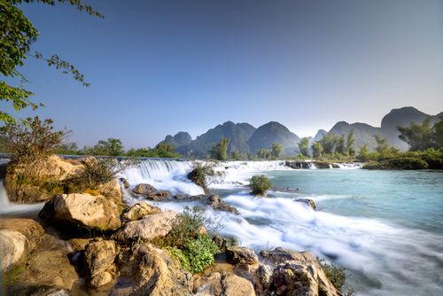 Free Landscape of small waterfall near rocky shore with green trees and grass near bushes and hills on background under blue cloudless sky in sunny summer weather Stock Photo
