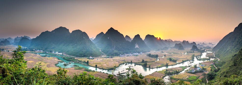Picturesque scenery of mountainous grassy valley with fields near river and green trees with plants at sunset in summer
