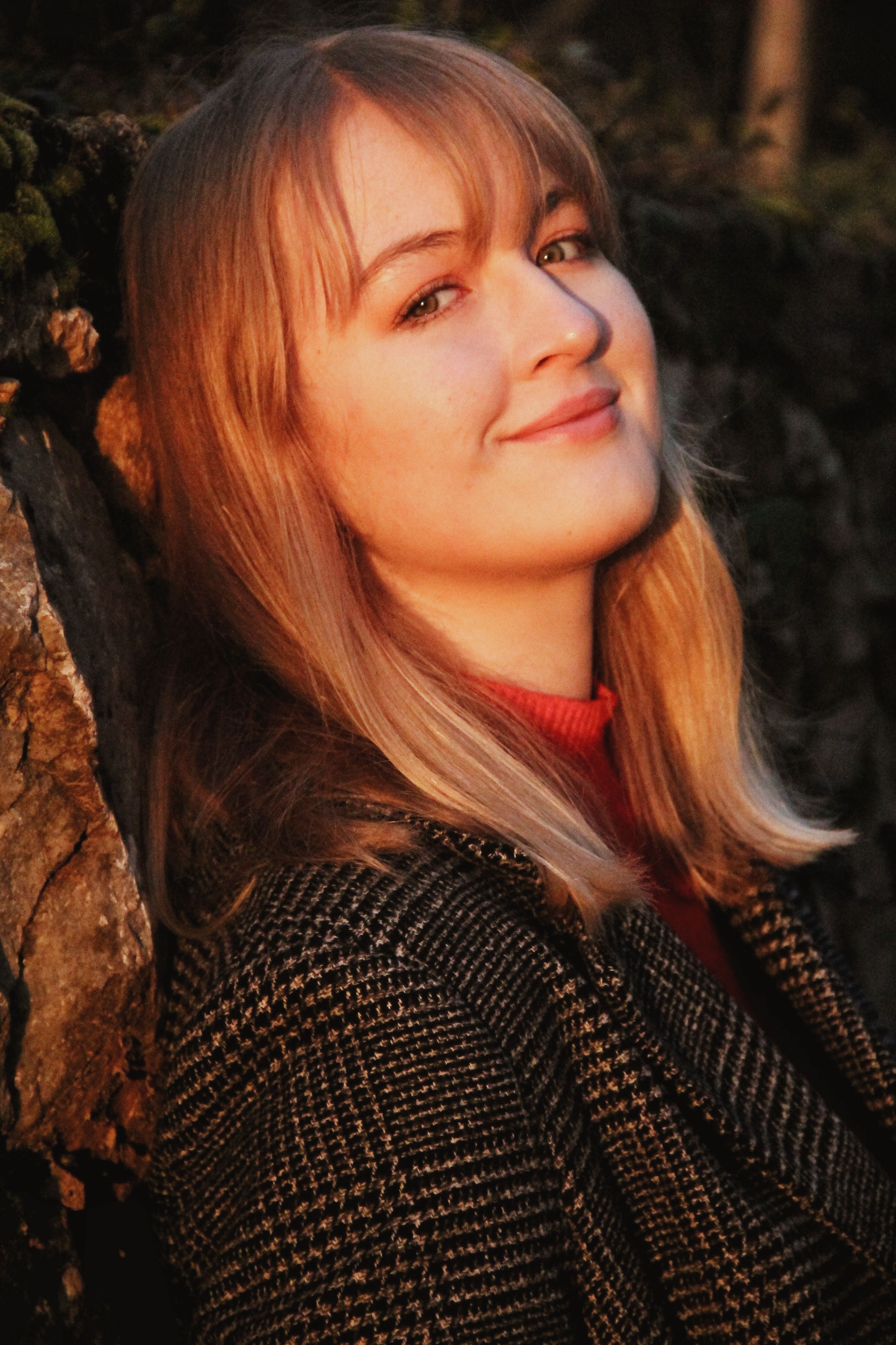 Close-up portrait of a smiling woman in a checkered coat leaning against a rock, enjoying natural surroundings.