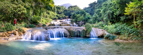 Wide angle of fast waterfalls streaming into calm river surrounded by green trees with lush vegetation and distant tourists in nature