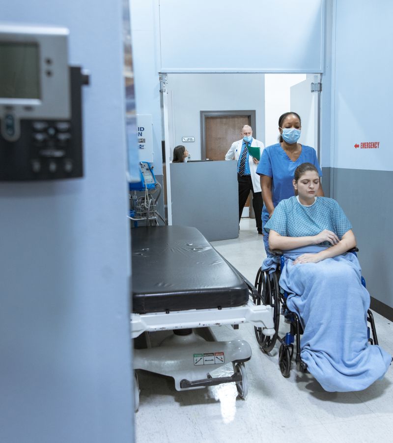 A nurse helps a patient in a wheelchair down a hospital corridor, reflecting care and medical professionalism.