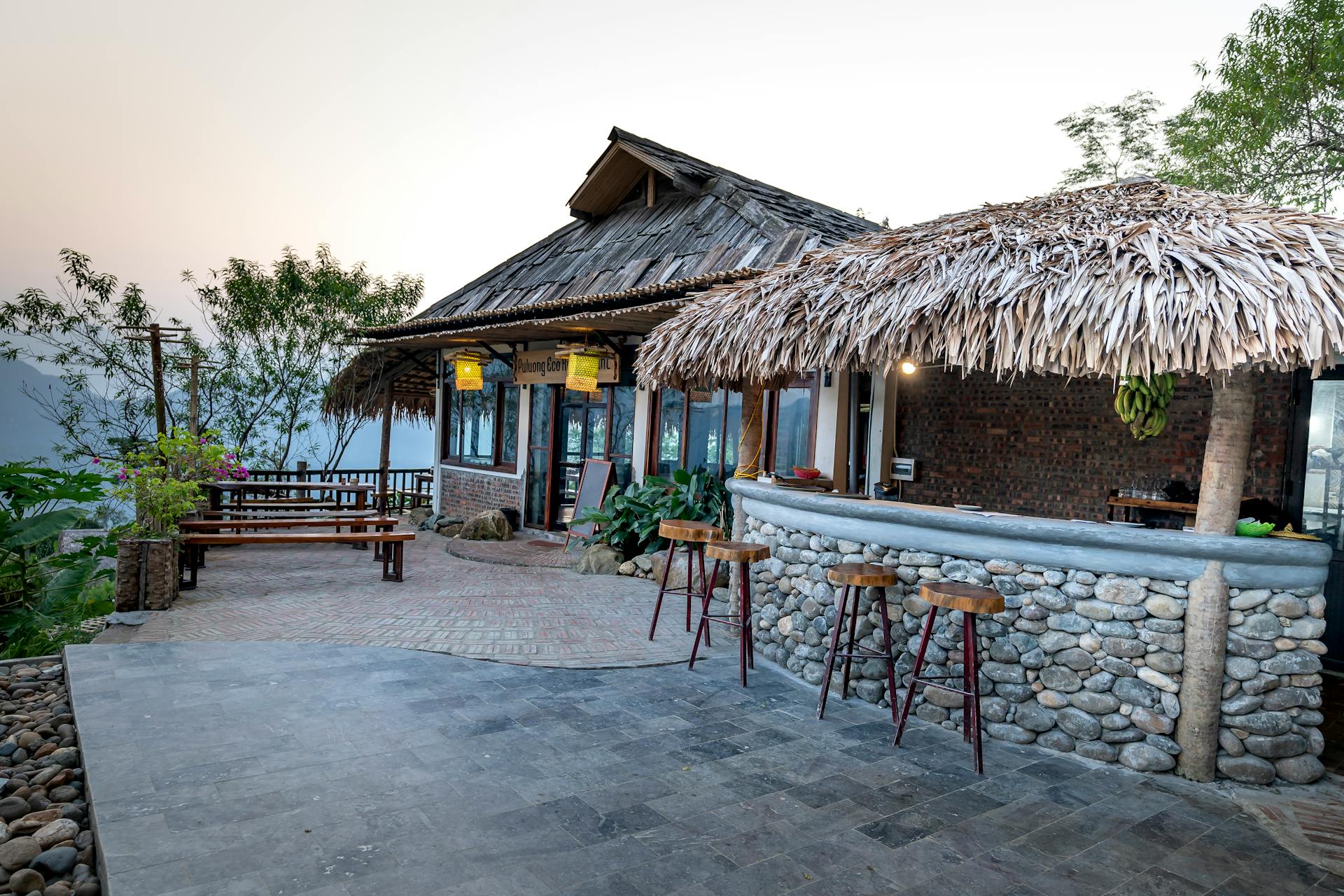 Stony bar with thatched roof and wooden villa located on paved veranda of tropical hotel in exotic country with green trees