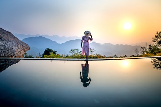 Back view of full body anonymous female in romantic dress and straw hat standing on poolside and looking away while enjoying sundown in amazing terrain