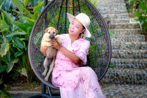 Satisfied mature ethnic lady in romantic dress and straw hat sitting in hanging chair and cuddling puppy in green garden