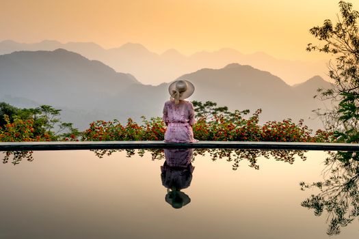 Back view of romantic unrecognizable woman in dress and straw hat sitting on poolside near smooth surface of water and looking away on sunset in mountain terrain