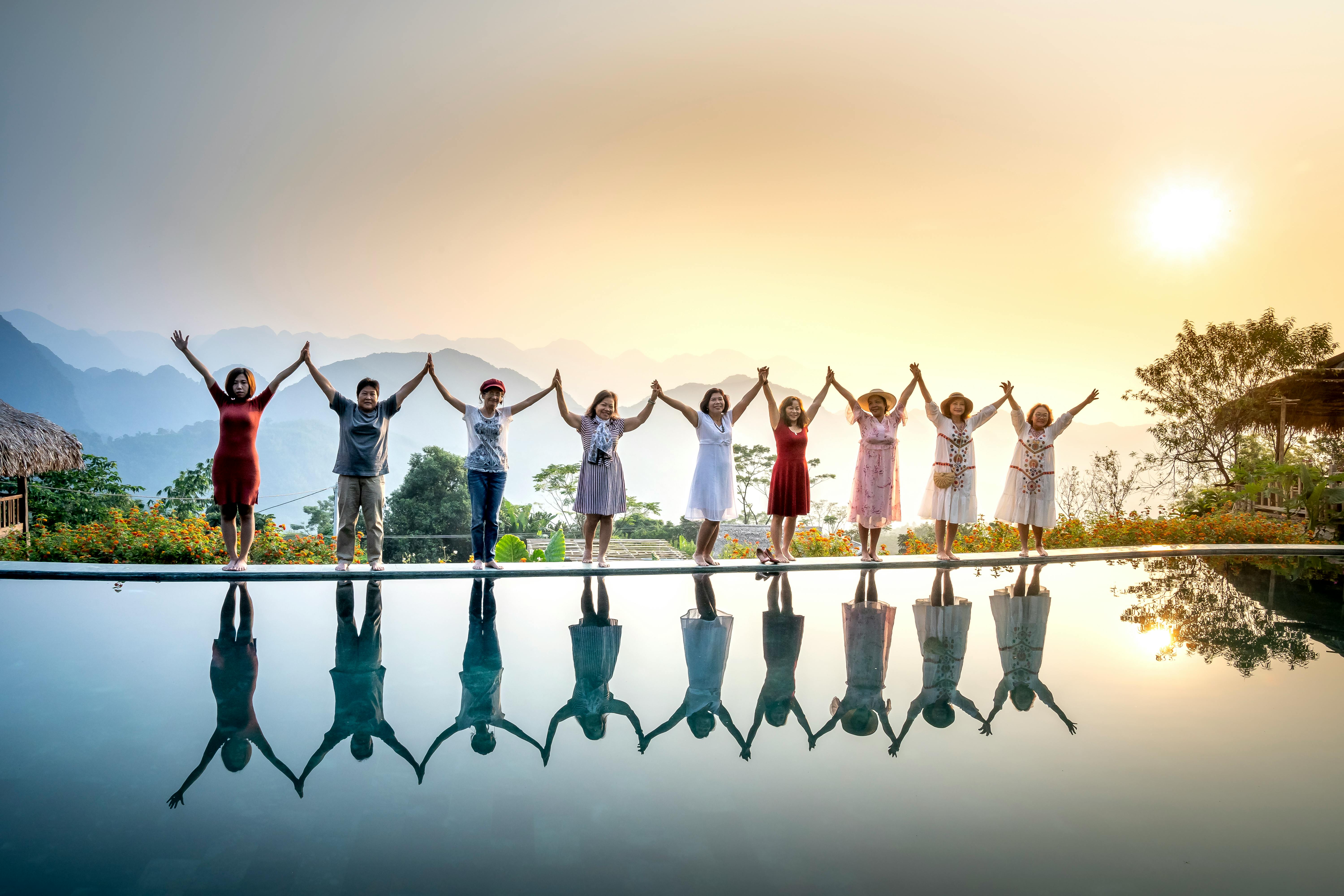 Happy full body ethnic people in summer clothes standing on poolside and holding hands with raised arms near calm water on sunset with amazing mountains on background