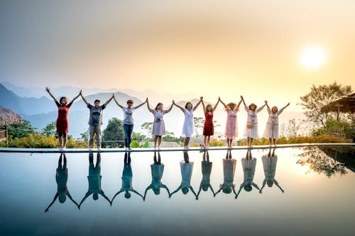 Free Happy full body ethnic people in summer clothes standing on poolside and holding hands with raised arms near calm water on sunset with amazing mountains on background Stock Photo