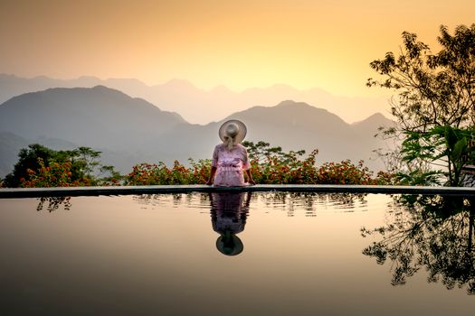 Back view of female in pink dress and hat sitting on poolside and admiring summer sunset in tropical terrain