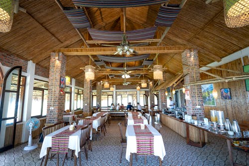 Various tables with colorful tablecloth and chairs in spacious cafeteria with dishes on counter and hanging lamps under wooden roof