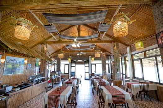 Rows of tables with chairs placed in cafeteria with hanging shining lamps and modern kitchenware on counter against opened door