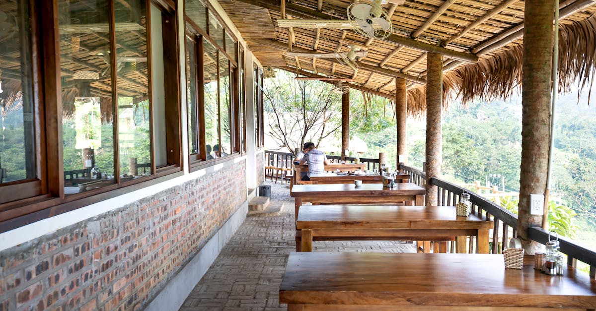 Photo by Quang Nguyen Vinh Wooden tables placed on veranda of cafeteria near windows and fence under wooden roof in tropical resort with green trees