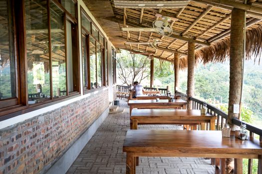 Wooden tables placed on veranda of cafeteria near windows and fence under wooden roof in tropical resort with green trees