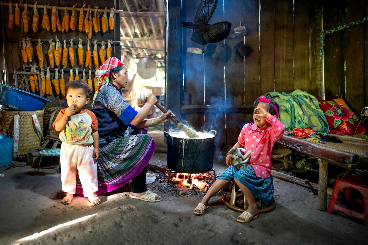 Side view of Asian woman in colorful outfit sitting with children and preparing food on bonfire in wooden rural barn in daytime