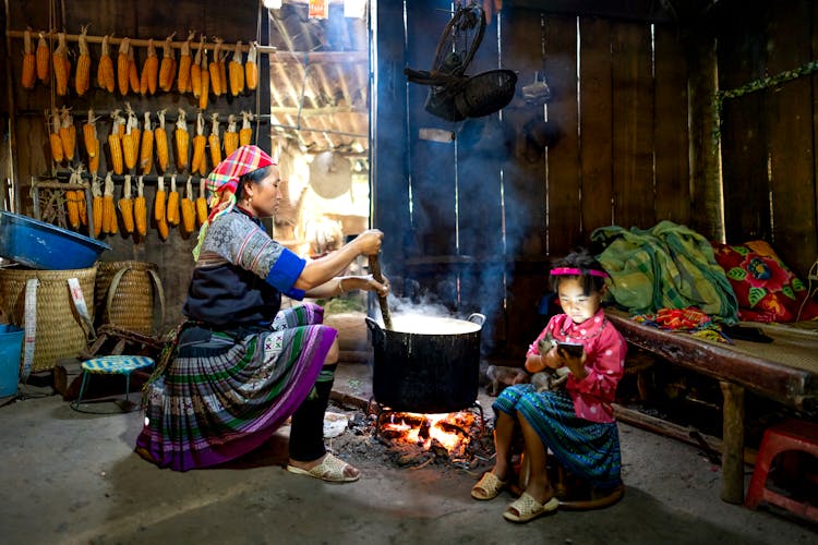 Ethnic Woman Preparing Food While Girl Using Smartphone In Wooden Barn