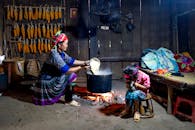 Content ethnic mom in traditional wear pouring rice into cauldron above fire against anonymous girl playing with cat at home