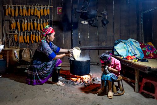 Free Content ethnic mom in traditional wear pouring rice into cauldron above fire against anonymous girl playing with cat at home Stock Photo