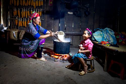 Free Smiling ethnic mom pouring rice into pot on fire against daughter sitting with cat on stool in poor house Stock Photo