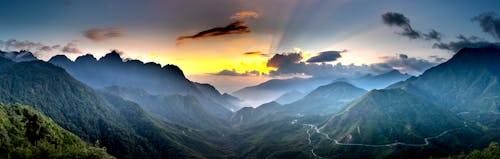 Panoramic view of majestic mounts with wavy roads under bright sky with fluffy clouds at sundown