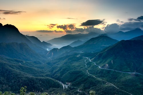 Scenic view of massive mountains with curved roadways under colorful cloudy sky at sundown in foggy weather