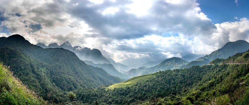 Wide angle of mounts with green trees under glowing sky with cumulus clouds in sunlight