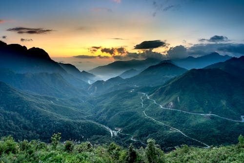 Spectacular view of magnificent mountains with lush green trees and wavy roadways under colorful sky with clouds at sundown