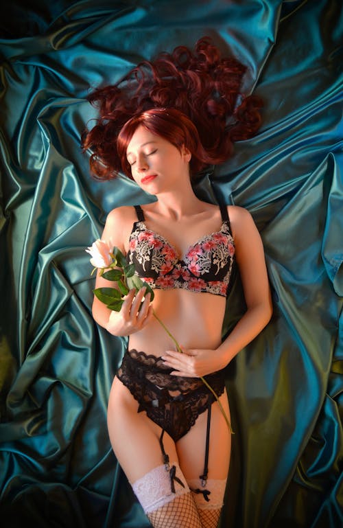 From above of relaxed young slim female with long red hair in sexy underwear and stockings holding fresh rose while lying on silk bed with closed eyes