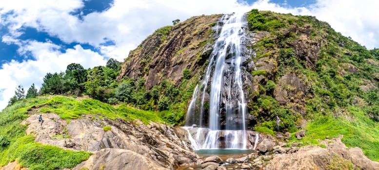 Panoramic view of a majestic waterfall cascading down a rocky cliff surrounded by lush greenery with a lone climber.