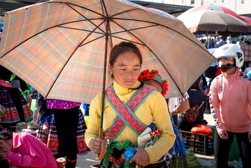 Pensive ethnic mother with umbrella carrying unrecognizable toddler girl against crop people while looking down in bazaar