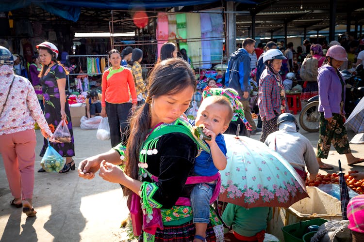 Ethnic Mother Carrying Baby With Treat In Local Market