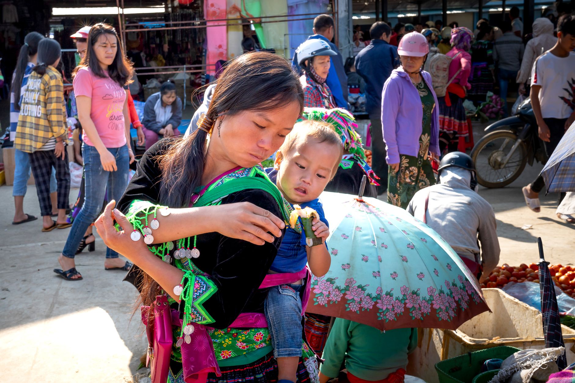 Ethnic mother with toddler child holding delicious sticky rice in banana leaf against unrecognizable people in urban bazaar