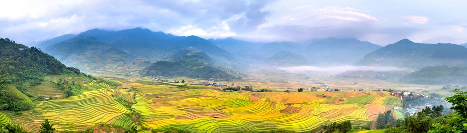 Scenery view of wavy agricultural fields against massive mountains with trees under cloudy sky in countryside in misty weather